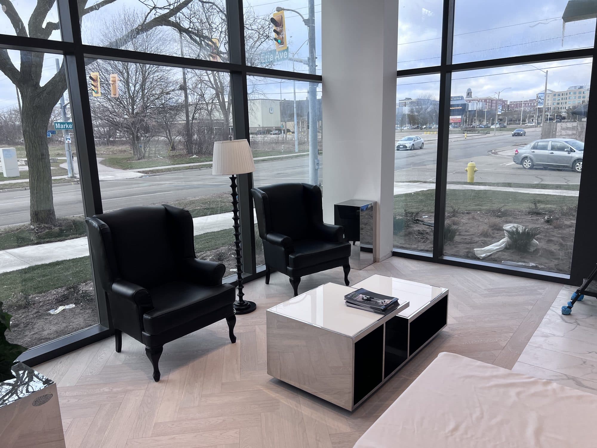 A ground-floor lounge in a new condominium building: two black wingback chairs and a low white table on a herringbone floor, behind glass running the full height of the wall, looking onto a Brantford street.