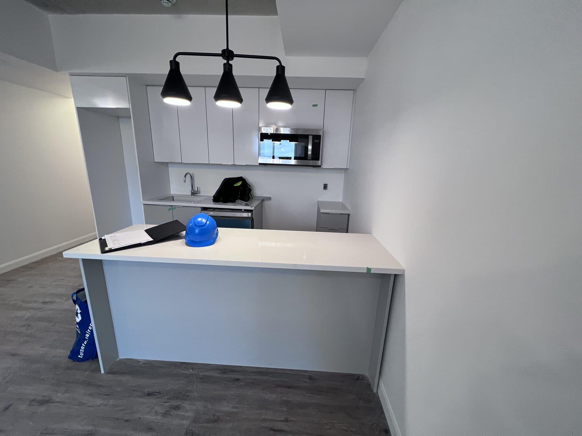 An unfinished condominium kitchen with white cabinets, a stainless microwave and three black pendant lights over a white island, a blue hard hat still sitting on the counter.
