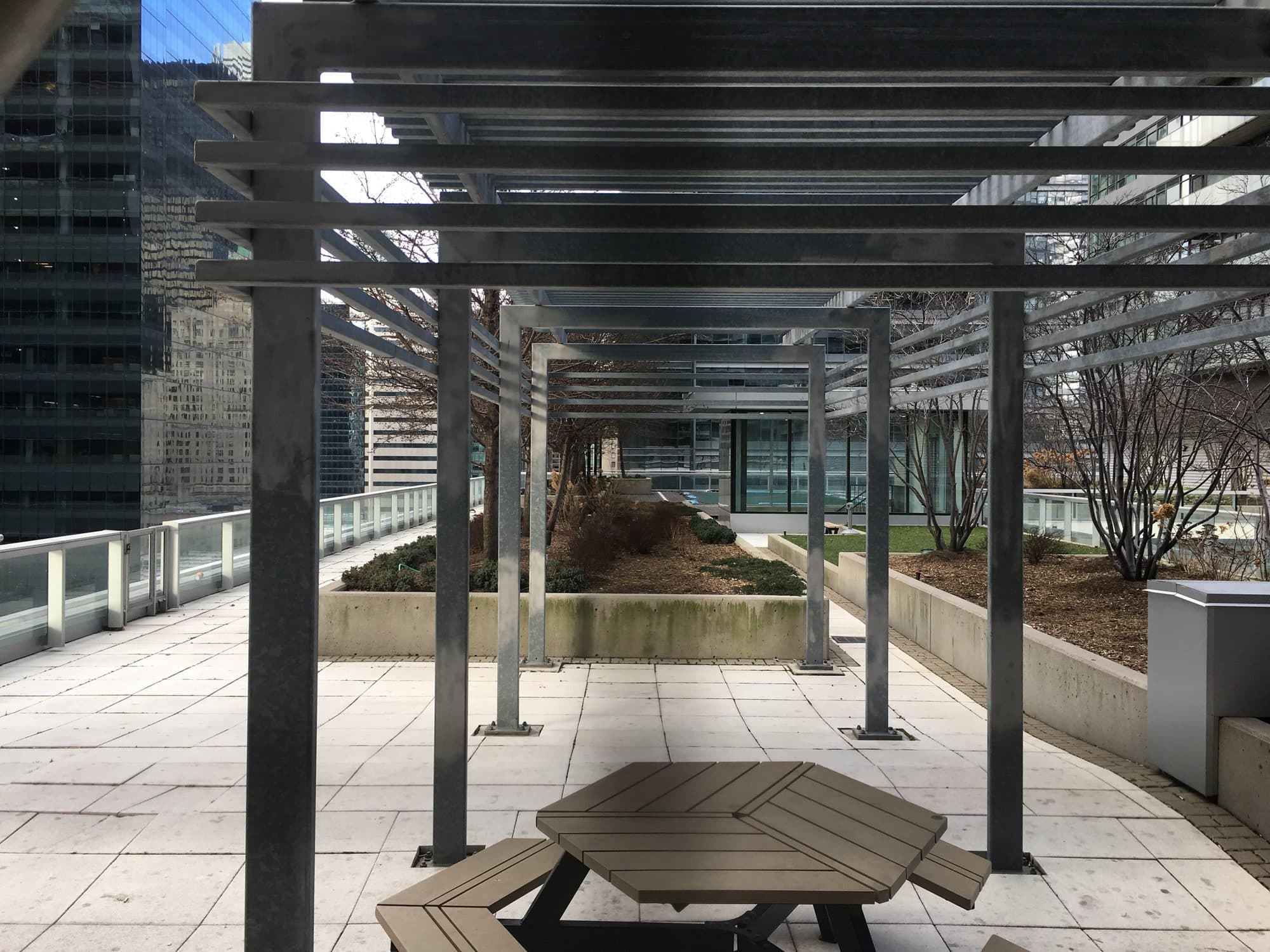 A shared rooftop terrace with a steel pergola and a wooden picnic table, enclosed on all sides by downtown Toronto office towers.