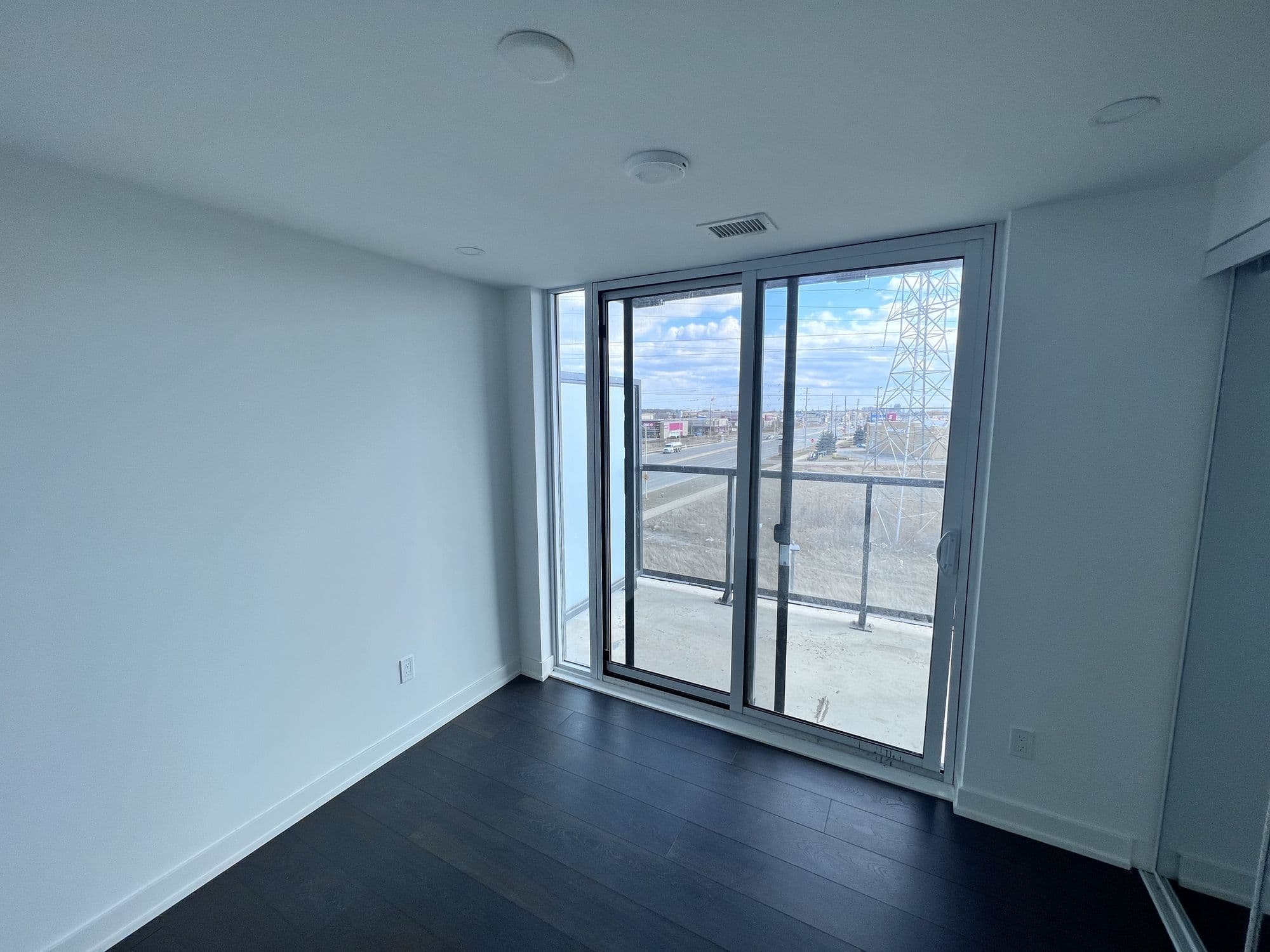 An empty condominium room with dark floors, bare white walls and a sliding balcony door looking out over rooftops.