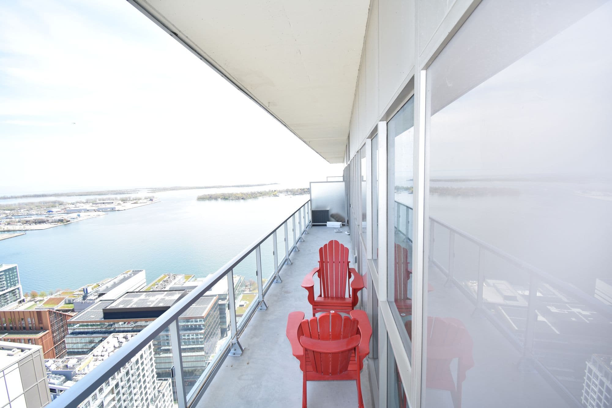 Two red Muskoka chairs on a high condominium balcony overlooking Toronto's harbour and the city skyline below.