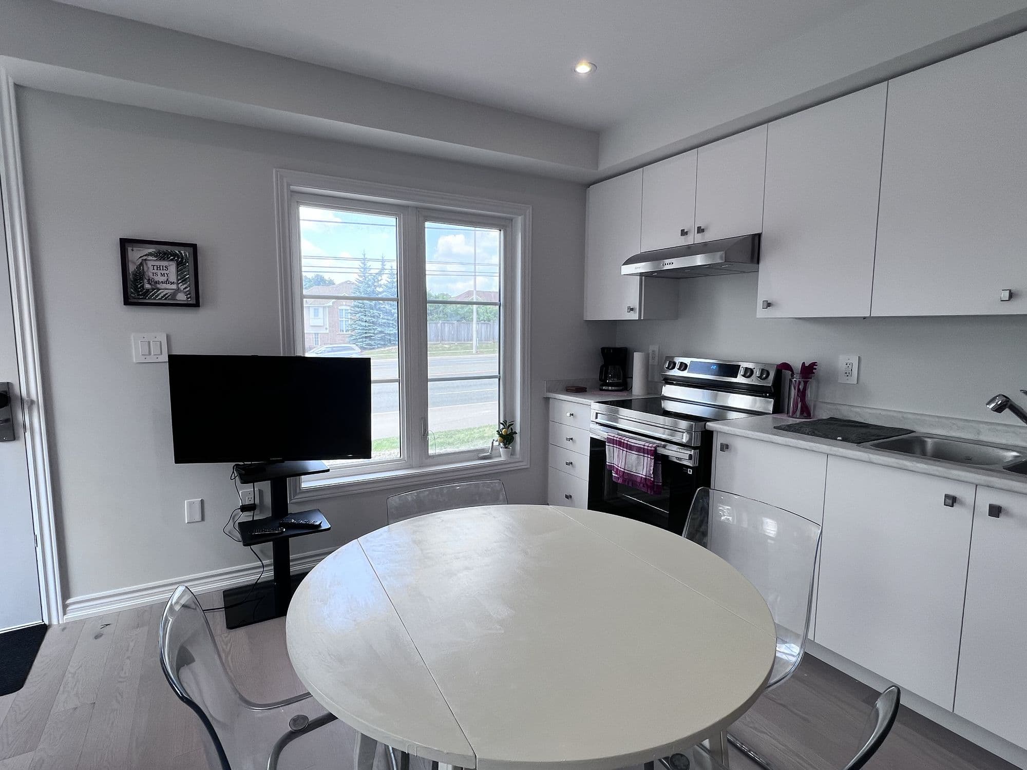 A bright furnished kitchen and dining area with white cabinets, a stainless range, a round table and a window onto the street.